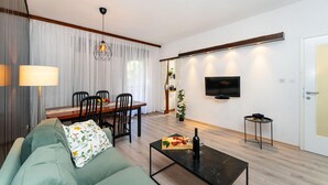 A modern living room with a sofa, a dining table with chairs, a coffee table with a bottle and glasses, a TV mounted on the wall, and a door with a handle.