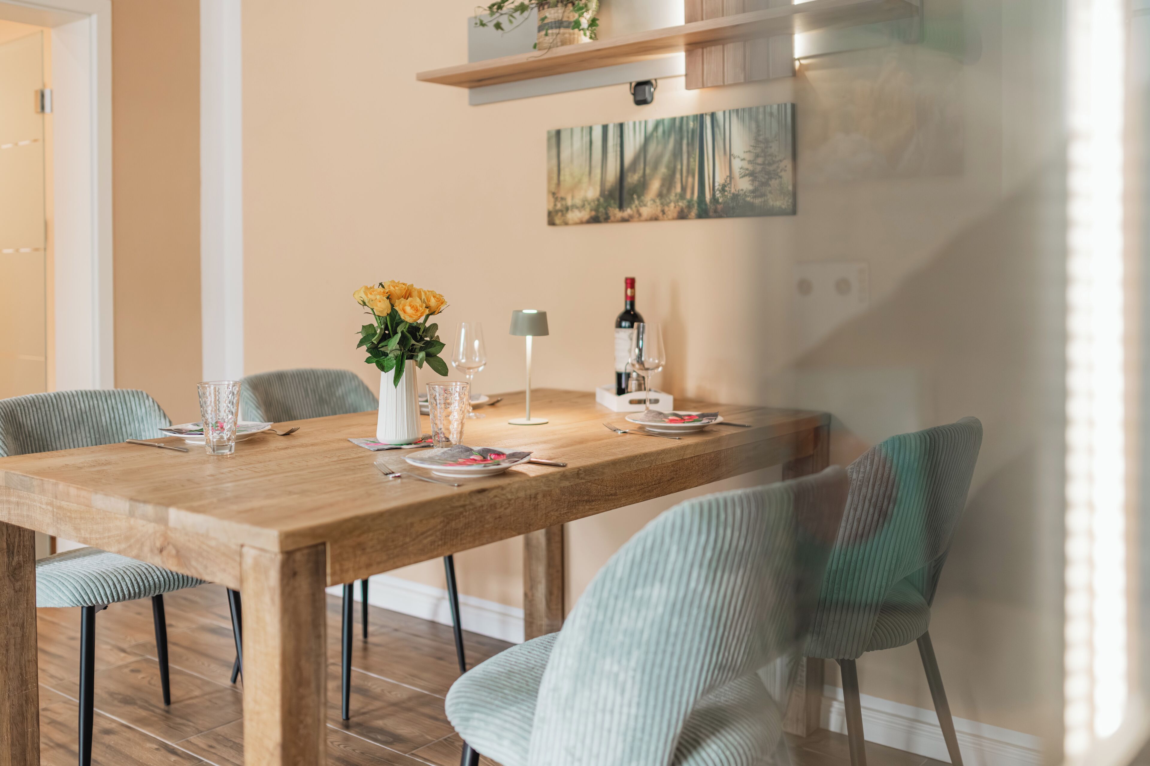 A dining area with a wooden table, chairs, a vase with flowers, wine glasses, and a bottle of wine.