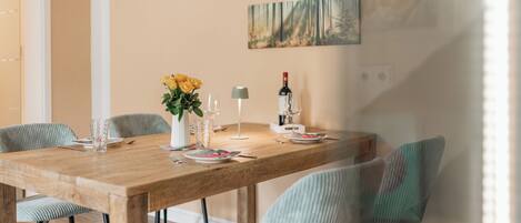 A dining area with a wooden table, chairs, a vase with flowers, wine glasses, and a bottle of wine.