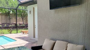 A sunlit patio area with a glass table, beige sofa, and a pool in the background.