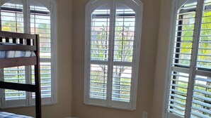 A bedroom with a bunk bed, a chair, and three windows with shutters.