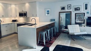 A modern kitchen with white cabinetry, a central island, and bar stools. There is a seating area with a white chair and a black ottoman.