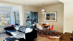 A modern living room with a black leather sofa, a dining table with red and white chairs, and a large window with a view of trees.