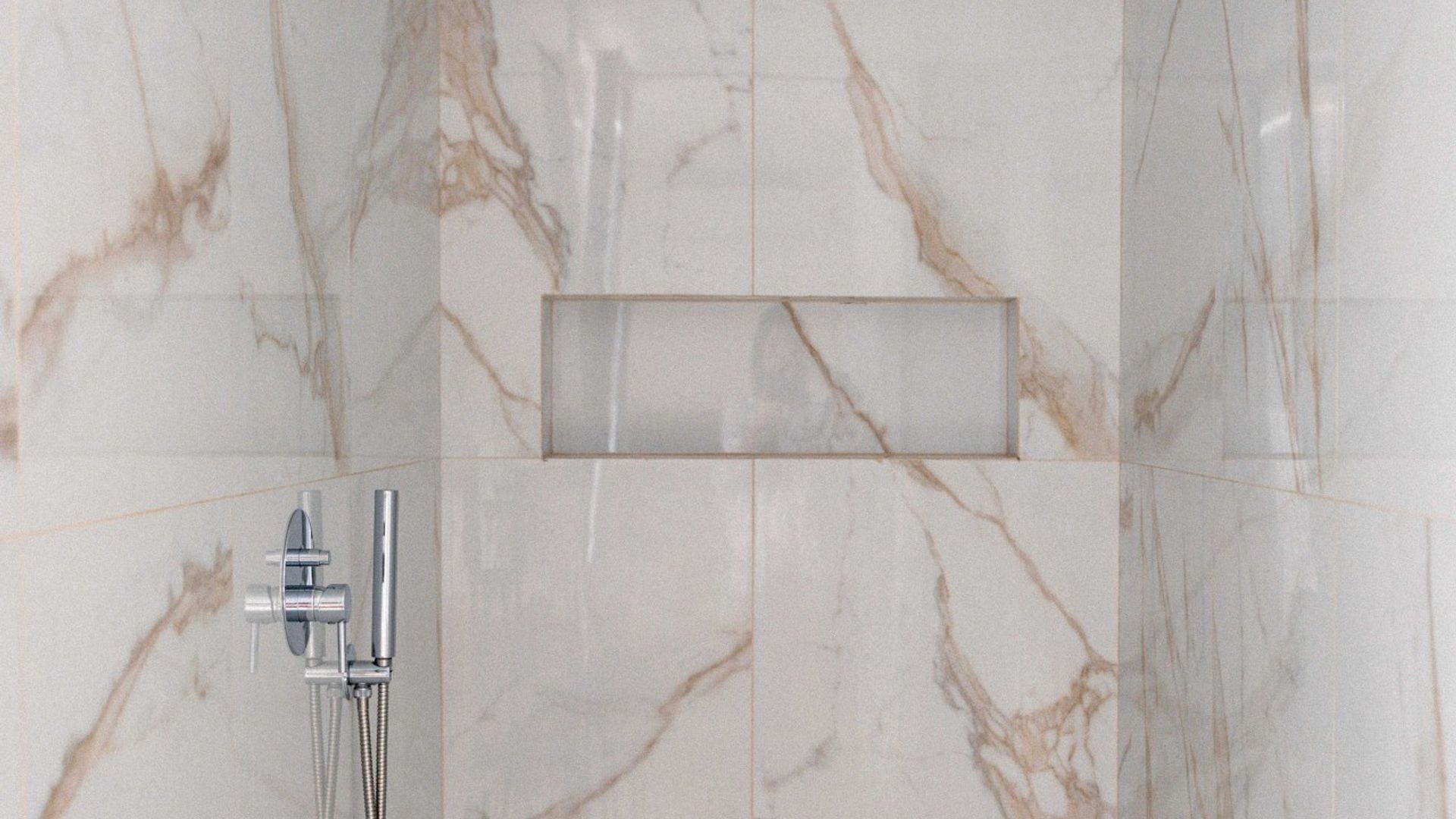 A bathroom with marble walls, a rectangular mirror, and a shower with a handheld showerhead.
