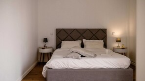 Uma cama bem arrumada com cabeceira, duas mesas de cabeceira com luminárias e um banquinho ao pé da cama.