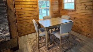Interior de una cabaña de madera con una mesa de comedor, cuatro sillas, una chimenea de piedra y dos ventanas.