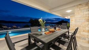 An outdoor dining area with a pool view, a stone wall, and a sunset in the background. - Awesome home in Posedarje (Posedarje)