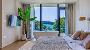 A modern bedroom with a large window, a bathtub, and a TV.