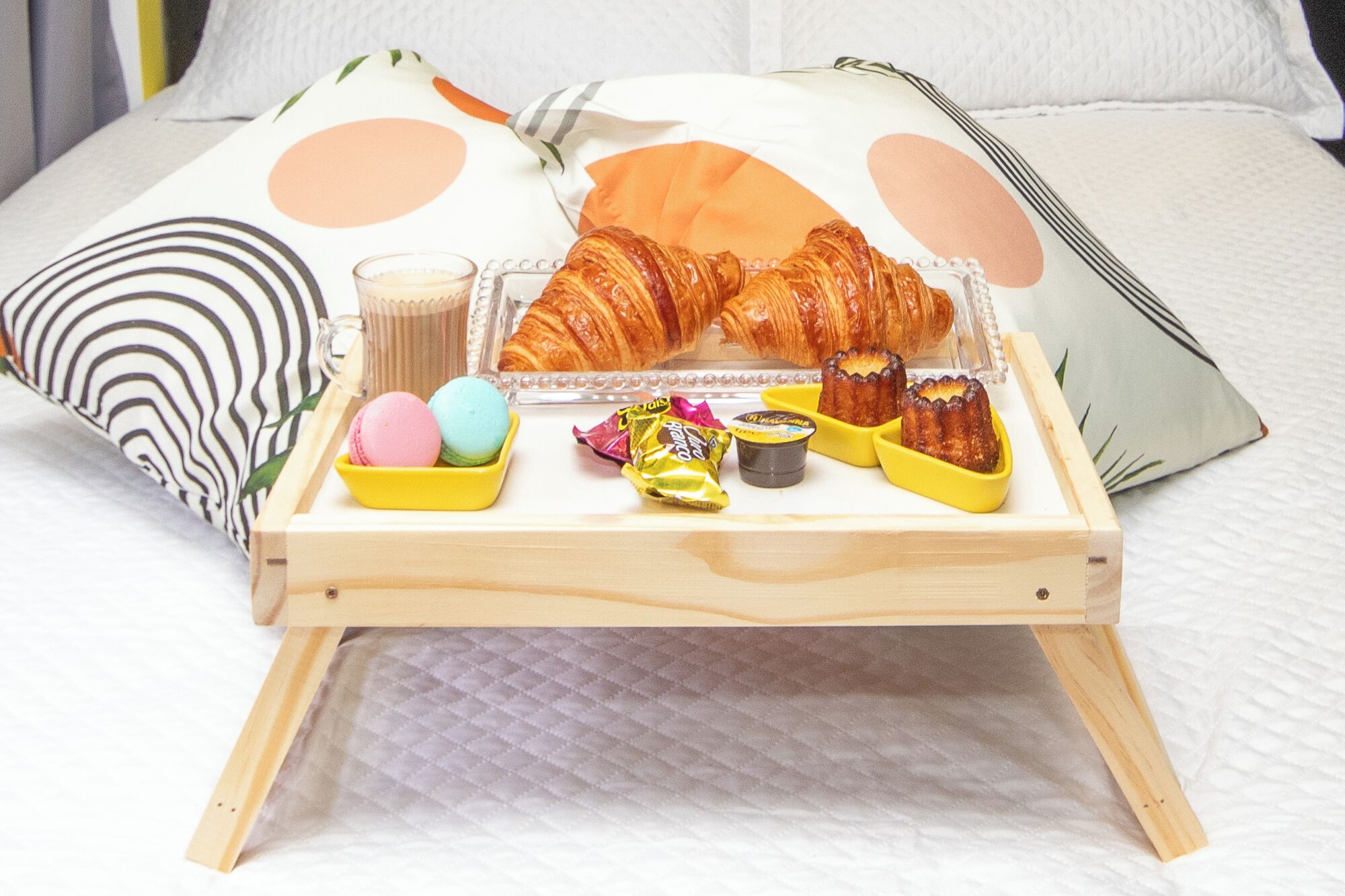 Una bandeja de madera con cruasanes, macarons y una taza de café sobre una cama con almohadones estampados.