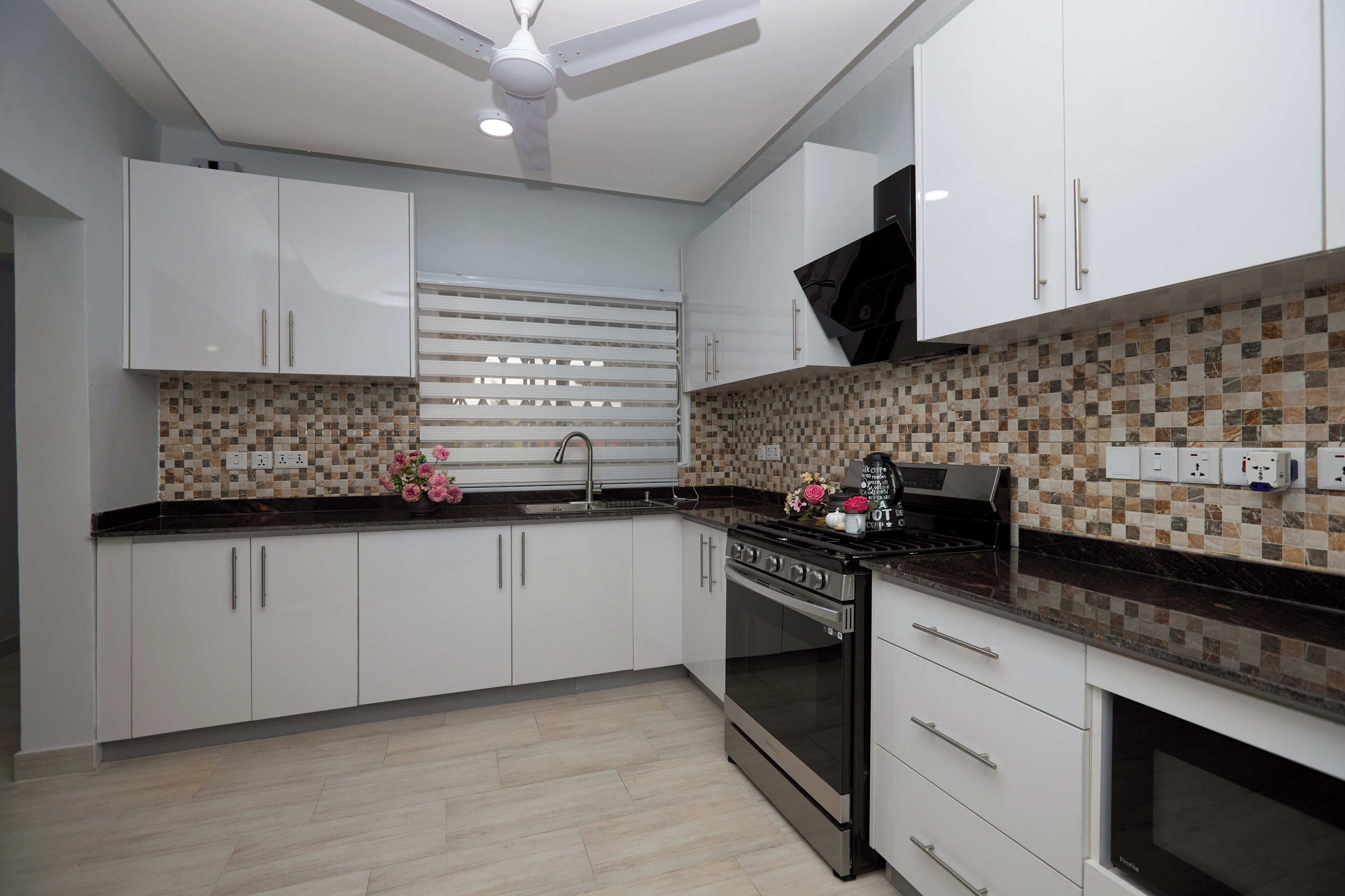 A modern kitchen with white cabinets, a black countertop, a tiled backsplash, a stove, and a ceiling fan.