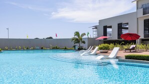 Una piscina con sillones de descanso y sombrillas, rodeada por un edificio de fachada blanca y una valla gris.