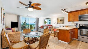 A spacious living area with a glass dining table, wicker chairs, a ceiling fan, and a kitchen with wooden cabinets and stainless steel appliances. - Beachfront Condo with Beach Access (Hauula)