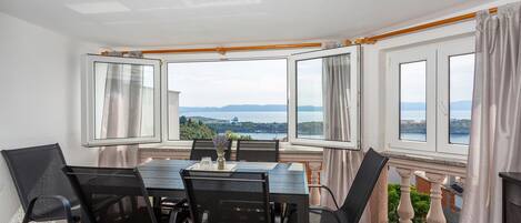 En matplats med ett bord och stolar, stora fönster med gardiner och utsikt över havet och kullarna.