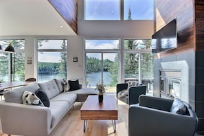 Un salon moderne comprenant un canapé, une table basse et des fauteuils, de grandes fenêtres offrant une vue sur une forêt et un lac, ainsi qu’une cheminée.