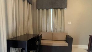 A compact living space with a dark-colored sofa, a matching dark-colored table, and a chair. The room has a tiled floor and is partially covered by beige curtains.