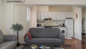 A compact living space with a grey sofa, a black coffee table with a bowl of grapes and a magazine, a small potted plant, and a kitchenette with white cabinets, a microwave, and a washing machine. - Golden Star AU – Elegance in Funchal (Funchal)