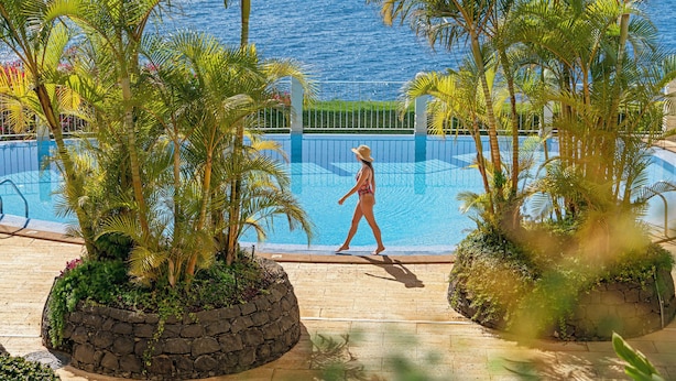 A pool area with a view of the ocean, surrounded by palm trees and a stone wall. - Arcadas III – Comfort and Sea View in Funchal (São Martinho)