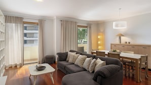 A modern living room with a grey sofa, a small white table, and a dining area with a wooden table and chairs. - Arcadas III – Comfort and Sea View in Funchal (São Martinho)