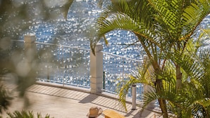 A sunlit outdoor lounge area with a person relaxing on a sunbed, surrounded by palm trees and a view of the sea. - Arcadas III – Comfort and Sea View in Funchal (São Martinho)