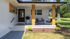 A house with a porch, wooden columns, a black door, and a small garden area with plants and a bench. - Private home near Greensboro GSO Airport 20% off in checkout limited time (Kernersville)
