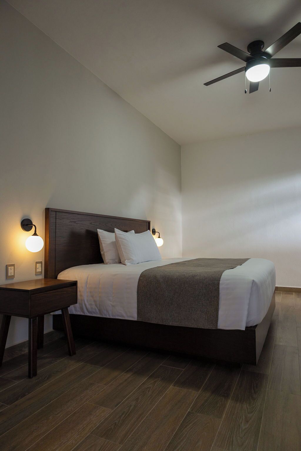 Un dormitorio moderno con una cama grande, cabecera de madera, dos mesitas de noche con lámparas de pared y un ventilador de techo.