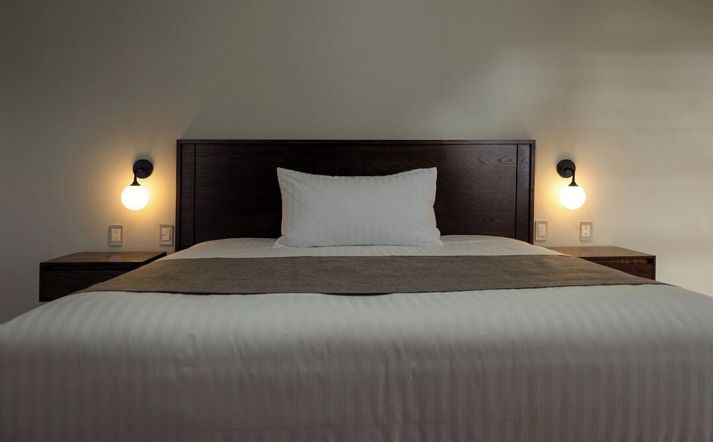 Una cama bien tendida con una almohada blanca y una manta marrón, flanqueada por dos mesitas de noche con lámparas de pared.