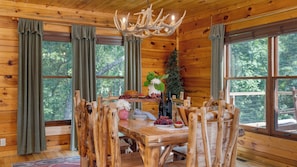 Interior de una cabaña de madera con una mesa de comedor, sillas, una araña y ventanas amplias.