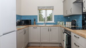 A modern kitchen with grey cabinets, a black microwave, a sink with a window above, and a stove. - Aunty Junes Cottage (Okehampton)