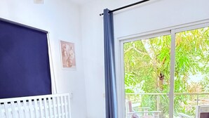 Et soveværelse med en seng, et vindue med gardiner, en hvid sengegavl og udsigt til grøn natur udenfor.