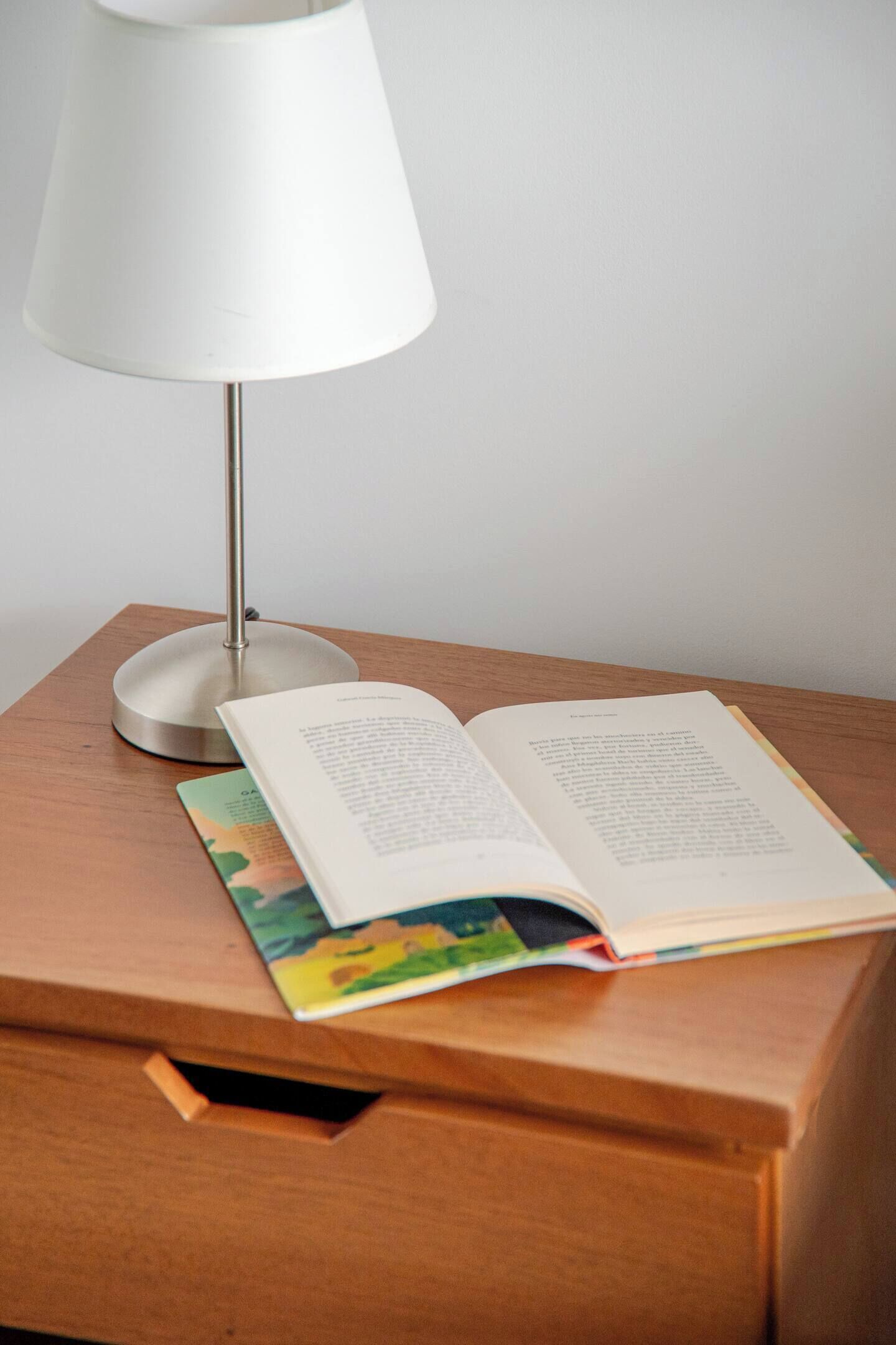 A wooden bedside table with a white lamp, an open book, and a colorful magazine.