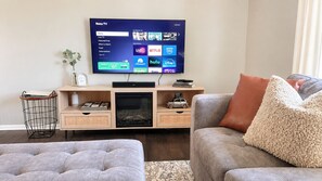 Olohuoneessa on litteänäyttöinen televisio, jossa näkyy Roku-televisiojärjestelmän käyttöliittymä, puinen viihdekeskus, jossa on pieni televisio ja pienoismalli autosta, sekä kaksi harmaata sohvaa, joissa on kuvioitu koristetyyny.