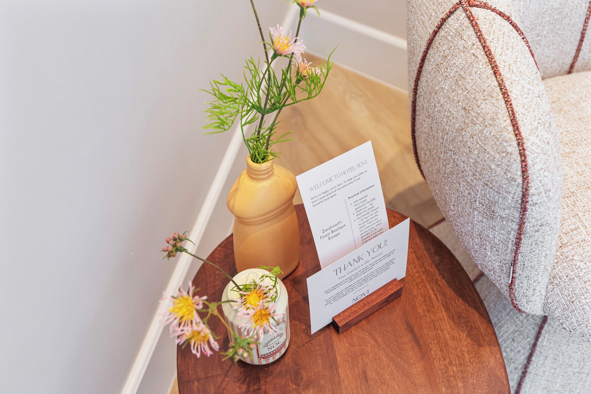 A wooden side table with a vase of flowers, a small bottle, and a welcome card.