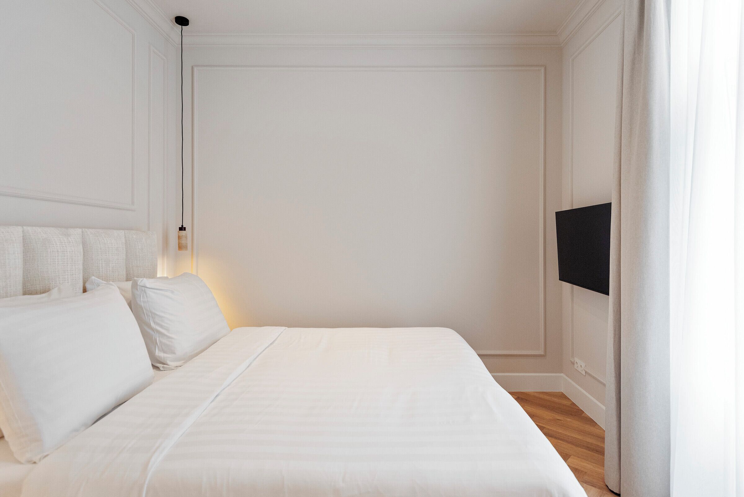 Una cama bien hecha con sábanas y almohadas blancas, una lámpara de noche y un televisor de pantalla plana montado en la pared.