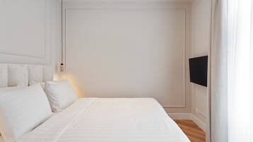 Una cama bien hecha con sábanas y almohadas blancas, una lámpara de noche y un televisor de pantalla plana montado en la pared.