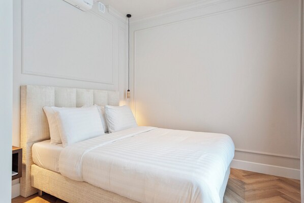 A neatly made bed with white linens in a room with a wooden floor and white walls.