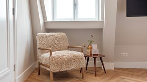 Una habitación con una butaca mullida, una pequeña mesa de madera con un florero y un menú, y una ventana con persianas blancas.