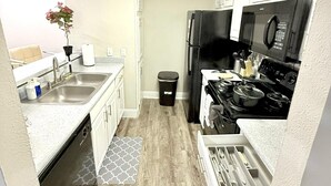 Una cocina compacta con armarios blancos, un refrigerador negro, un microondas y una estufa. Hay un pequeño cubo de basura y una planta en maceta sobre la encimera.