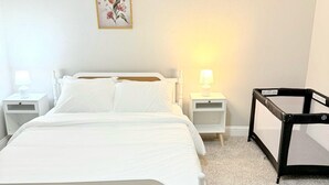 Un dormitorio con una cama blanca, dos mesitas de noche con lámparas, un cuadro colgado en la pared y una pequeña mesa con una pantalla.