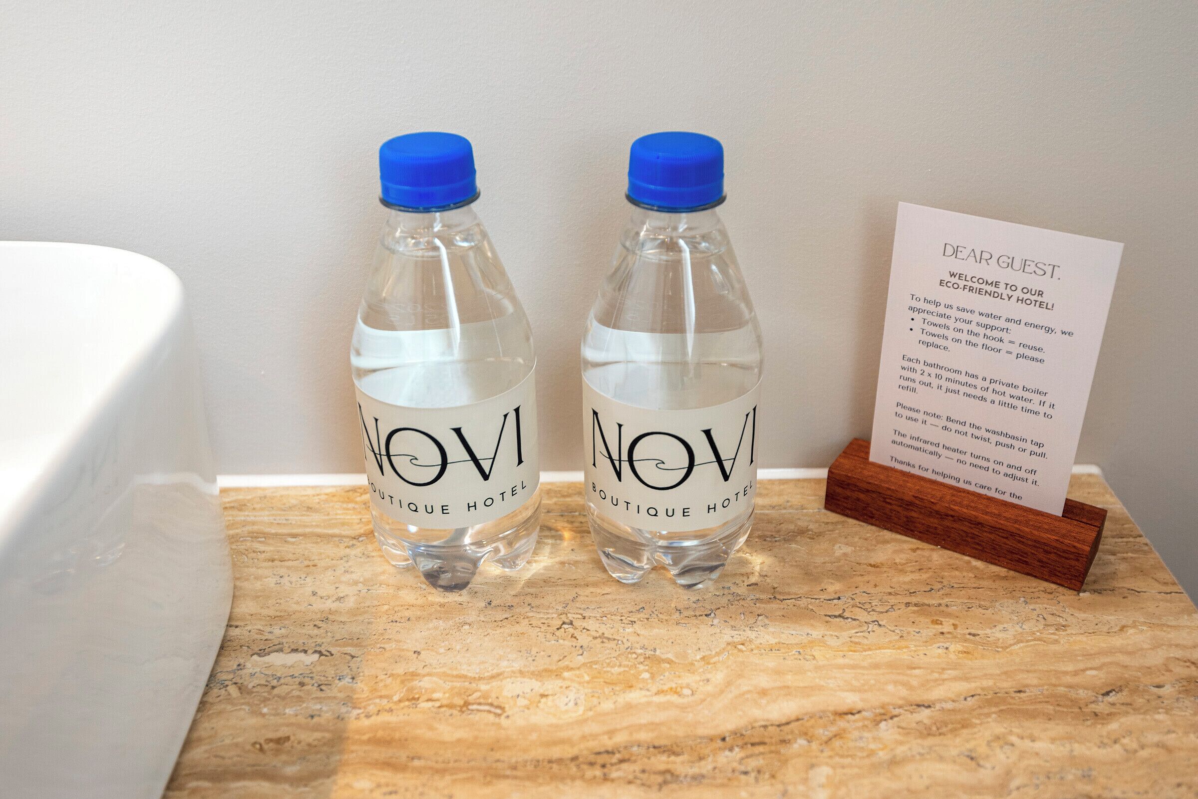 Two water bottles labeled "NOVI Boutique Hotel" on a marble countertop next to a white bathtub and a welcome sign.