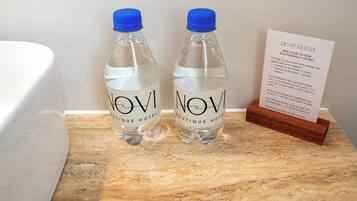 Two water bottles labeled "NOVI Boutique Hotel" on a marble countertop next to a white bathtub and a welcome sign.