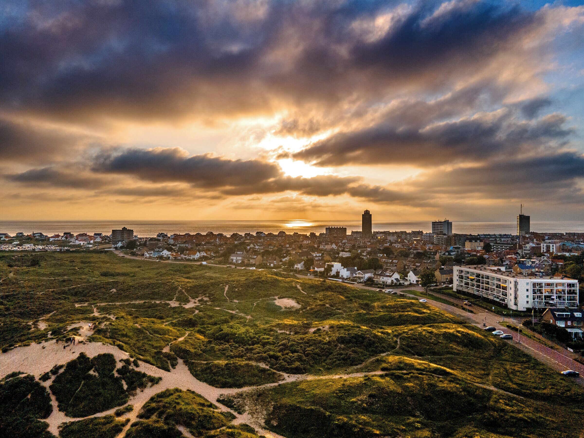 Un paysage urbain côtier, avec un mélange de bâtiments résidentiels et commerciaux, une tour imposante et un coucher de soleil sur l’océan.
