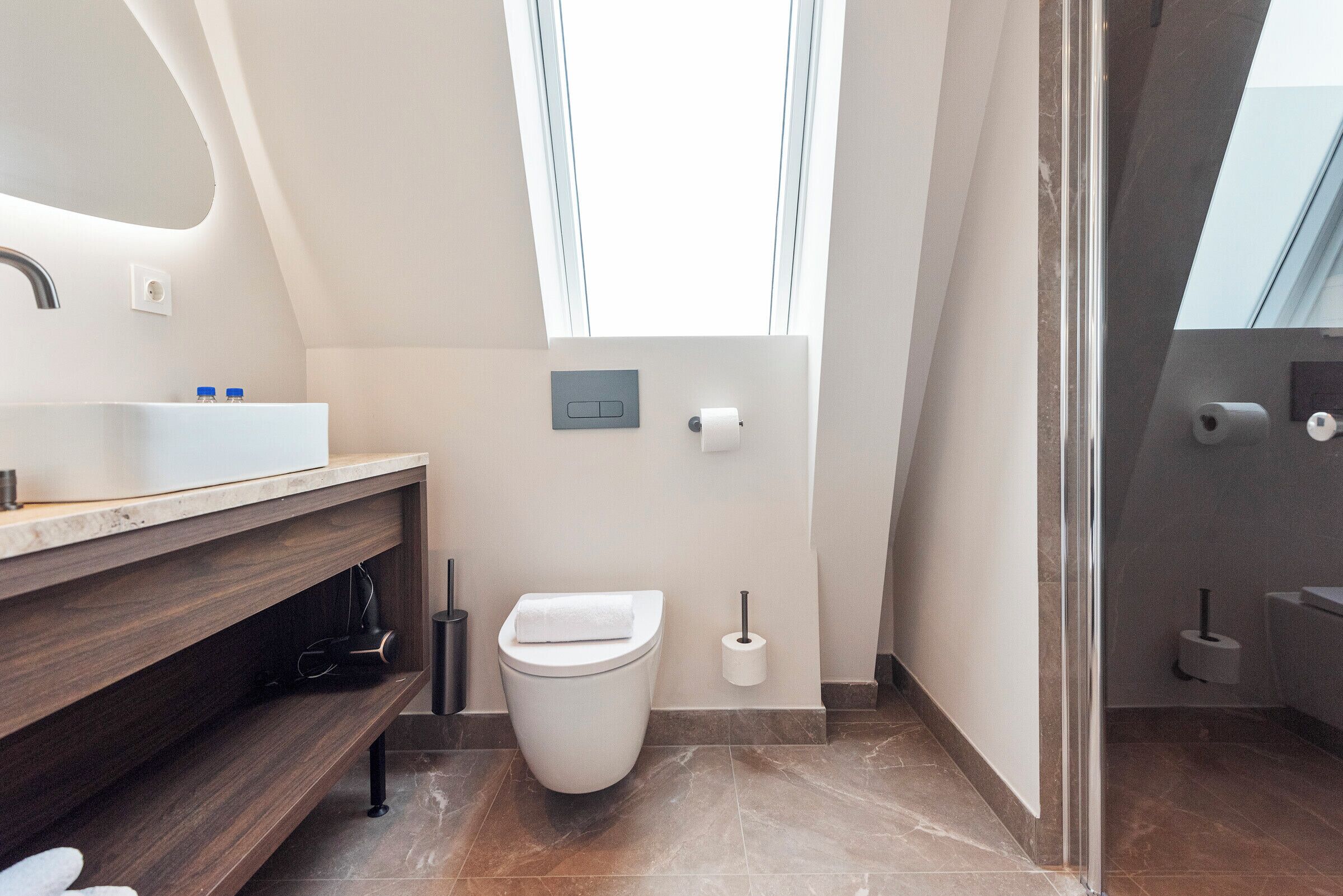 Un baño moderno con inodoro blanco, lavamanos con tocador de madera y ventana que proporciona luz natural.