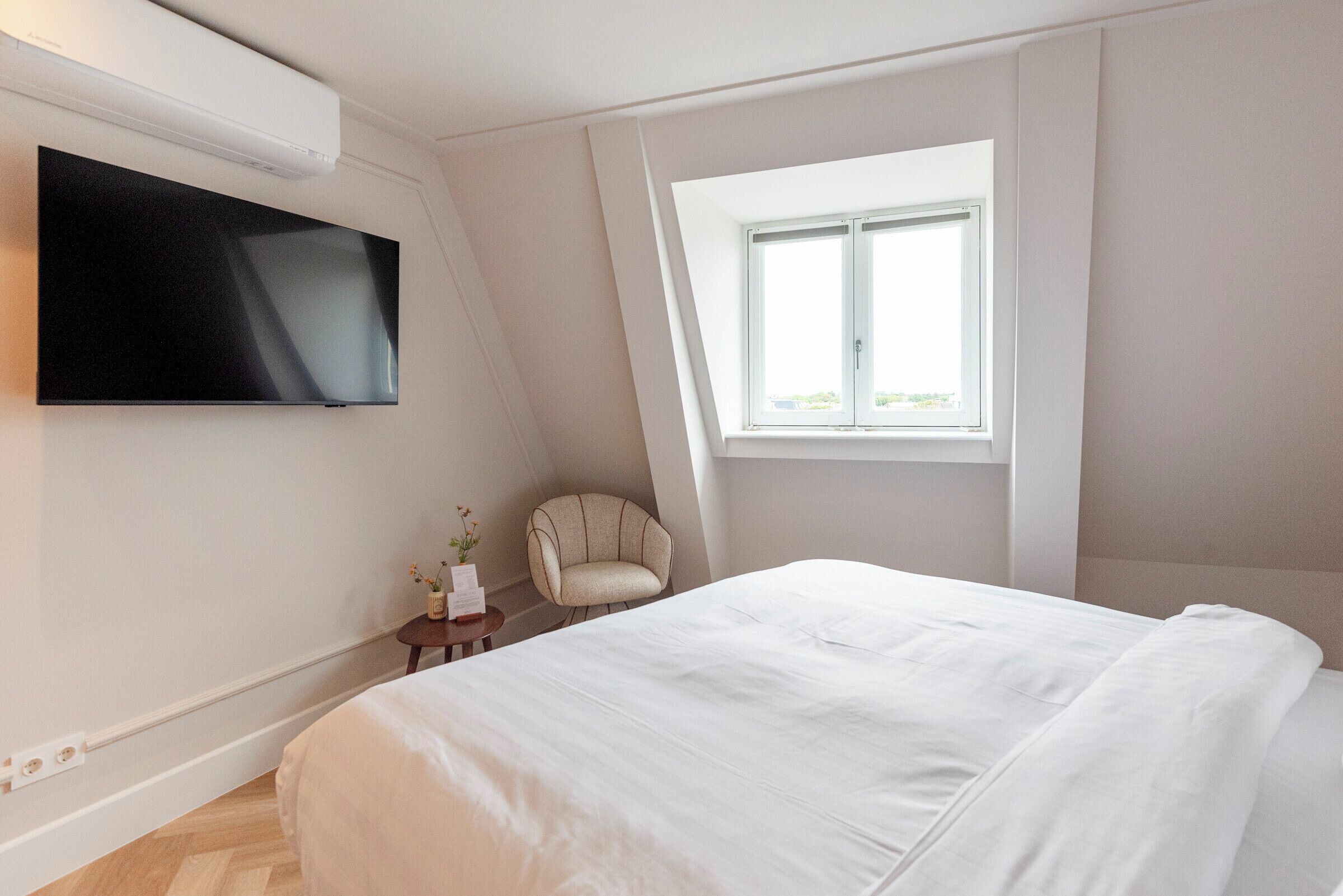 Ein Schlafzimmer mit einem großen Bett, einem flachbildfernseher an der Wand, einem kleinen Nachttisch mit Blumen, einem Sessel und einem Fenster mit Blick ins Freie.