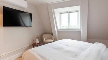 Ein Schlafzimmer mit einem großen Bett, einem flachbildfernseher an der Wand, einem kleinen Nachttisch mit Blumen, einem Sessel und einem Fenster mit Blick ins Freie.