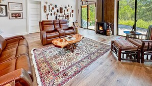 Ein geräumiges Wohnzimmer mit einer Ledersofa, einem Holztisch und einem großen Teppich. Ein Kamin und ein Blick ins Freie durch große Fenster sind vorhanden.