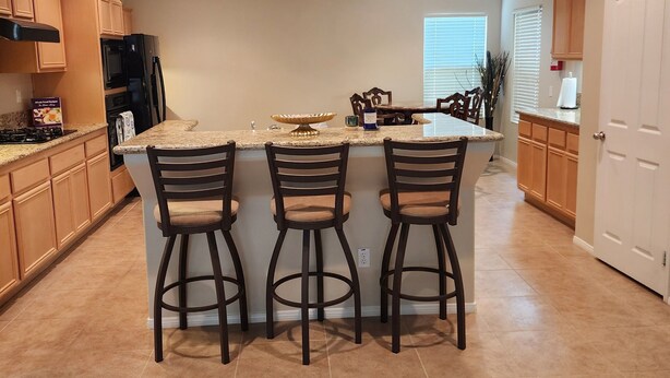 A kitchen with a central island, three bar stools, wooden cabinets, a black refrigerator, and a window with blinds.