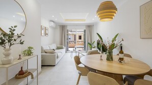 Una sala de estar moderna con sofá, mesa de comedor y sillas. Hay un espejo grande en la pared y una lámpara colgante.