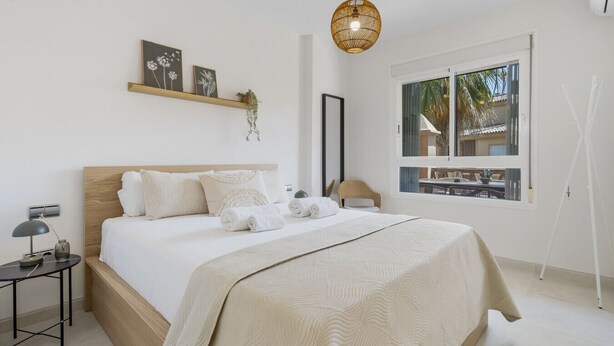 Un dormitorio moderno con una cama grande, una mesita de noche, una lámpara colgante y una ventana con vistas a palmeras.