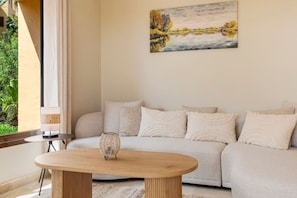 Ein Wohnzimmer mit einem beigefarbenen Sofa, einem runden Couchtisch, einem Bild an der Wand und Blick auf Grünfläche draußen.