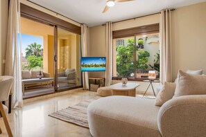 Een ruime woonkamer met een grote televisie, een bank, een eettafel en uitzicht op een tropische tuin.